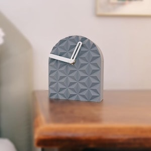 Pode incluir: Um relógio de mesa cinza em forma de arco com um padrão geométrico triangular na superfície frontal. O relógio possui dois ponteiros brancos simples posicionados no centro, indicando as horas sem números ou marcações no mostrador.