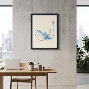 May include: A framed wall art print featuring a blue abstract line drawing on a cream background. The artwork is enclosed in a simple black frame and displays a series of fluid, overlapping blue strokes that form a central, organic shape.