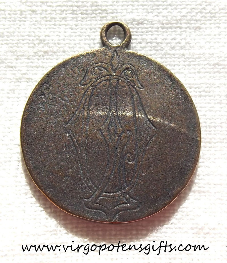 May include: A round, bronze-colored pendant with a decorative design. The pendant has a loop at the top for hanging.
