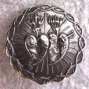 Könnte beinhalten: Ein silberfarbener Anhänger aus Metall mit zwei ineinander verschlungenen Herzen, die von Pfeilen durchbohrt werden, umgeben von einem Dornenkranz. Der Anhänger hat eine strukturierte Oberfläche und ein detailliertes Design.