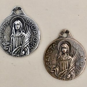 May include: Two silver-toned metal medallions with a raised image of a woman holding a chalice and a palm branch. The medallions are round and have the words "SANCTA LUCIA ORN" inscribed around the edge.