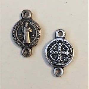 May include: Two silver-toned metal charms with a loop on each side. One charm features a depiction of a saint with a staff and a halo. The other charm features a cross with a circle around it. Both charms have text around the edges.