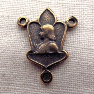 May include: A bronze-colored metal charm with a detailed image of a person's profile. The charm has two loops on the top and one loop on the bottom, suggesting it is a religious charm or a part of a rosary.