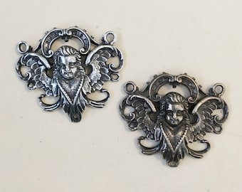 Puede incluir: Dos adornos antiguos de metal plateado con forma de querubín. Cada querubín tiene alas detalladas, una cara serena y adornos decorativos. Cuentan con pequeños lazos para colgar y un agujero en la parte inferior.