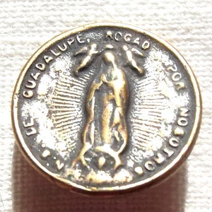 May include: A silver and gold metal religious medal with a depiction of the Virgin Mary and the text "DE GUADALUPE, REGO POR NO OTRO" around the edge.