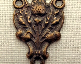 May include: A brass-colored metal charm with a detailed design of a thistle flower. The charm has two holes at the top and one at the bottom.