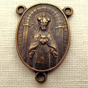 May include: An oval, bronze-coloured religious pendant. The pendant features a detailed relief of a crowned figure with a heart, surrounded by a halo and architectural elements. It has three holes for attachment.