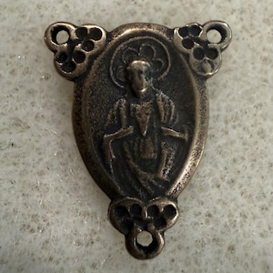 May include: A bronze-toned metal rosary centerpiece featuring a central oval relief of a figure with a halo. The piece has a triangular shape with decorative floral-like cutouts at each of the three corners, each containing a small circular hole.
