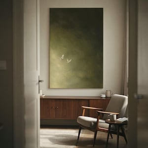 May include: A large rectangular wall art piece featuring a textured, deep olive green background with two small, white birds flying in the lower left quadrant. The painting is displayed on a wall above a wooden cabinet and an armchair.