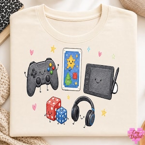 Puede incluir: Una camiseta de color crema con un estampado gráfico de varios artículos de juego y creativos con caras sonrientes, incluyendo un mando de juego negro, una tableta con lápiz óptico, auriculares, dos dados y una tarjeta rectangular de pixel art.