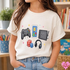 May include: A cream-colored t-shirt featuring a graphic print of various gaming items with smiling faces, including a black game controller, a tablet with a stylus, a pair of headphones, and two dice, all surrounded by small colorful hearts and stars.