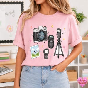 Peut inclure: Un t-shirt rose clair avec une impression graphique représentant un appareil photo, un objectif, un flash, un trépied et une photo encadrée de style dessin animé. Chaque élément a un visage souriant, agrémenté de petits cœurs et étoiles.
