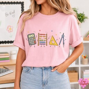 May include: A light pink short-sleeve t-shirt featuring a graphic print of four anthropomorphic school supplies: a grey calculator, a wooden abacus with colourful beads, a yellow triangle ruler, and a grey compass, all with smiling faces.