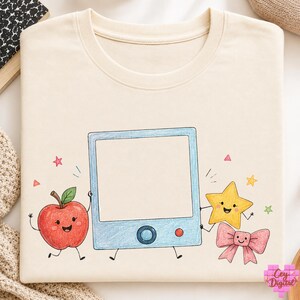 May include: A cream-coloured t-shirt featuring a central graphic of a light blue square frame with a red button and a blue dial. The frame is flanked by a smiling red apple, a yellow star, and a pink bow, all with hand-drawn pencil textures.