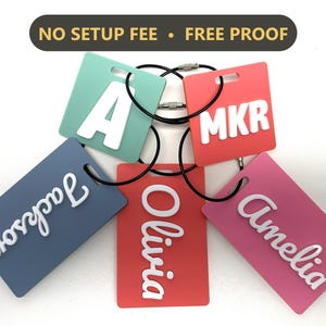 May include: Five rectangular luggage tags in mint, coral, blue, red, and pink with white raised lettering reading A, MKR, Jackson, Olivia, and Amelia. Each tag features a black circular wire loop for attachment and text NO SETUP FEE • FREE PROOF.