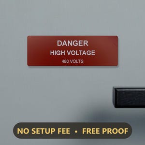 May include: A rectangular red safety sign with white text that reads DANGER HIGH VOLTAGE 480 VOLTS. Below the sign, a dark rounded rectangular label features the yellow text NO SETUP FEE • FREE PROOF. A black handle is visible on the right.