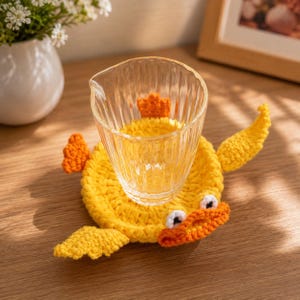 May include: A clear, vertically ribbed glass pitcher sits atop a yellow crocheted coaster shaped like a duck. The coaster features orange wings, a tail, and a beak, with two small white and black eyes positioned above the orange beak.