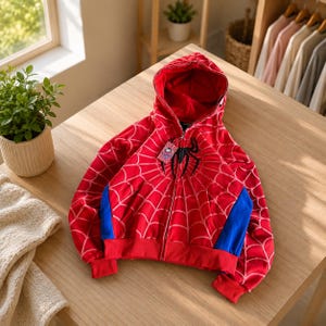 May include: A red zip-up hoodie featuring a black spider emblem on the chest and a white spiderweb pattern across the fabric. The garment includes a hood, long sleeves with blue side panels, and a small rectangular tag attached to the zipper.