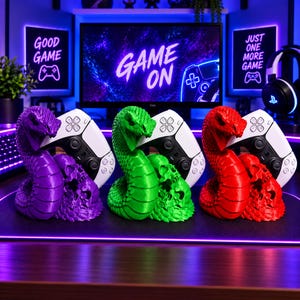 May include: Three 3D-printed controller stands in purple, green, and red, each featuring a coiled snake and a skull base. These stands hold white and black game controllers, positioned in front of a monitor displaying the text GAME ON.