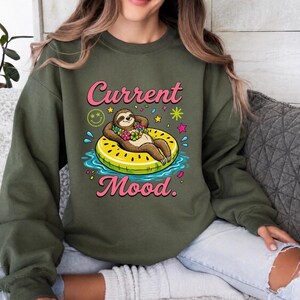 May include: An olive green crewneck sweatshirt featuring a graphic of a brown sloth wearing a floral lei, lounging on a yellow watermelon-patterned pool float in blue water. The text Current Mood is printed in pink above and below the illustration.