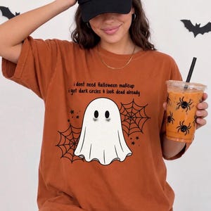 Pode incluir: Uma t-shirt cor de laranja queimado com um gráfico de um fantasma branco com olheiras, teias de aranha pretas e o texto: i dont need Halloween makeup i got dark circles & look dead already. A t-shirt tem mangas curtas e gola redonda.