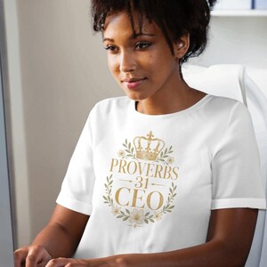 Puede incluir: Una camiseta blanca de manga corta con un estampado gráfico de color dorado. El diseño incluye una corona en la parte superior, el texto "PROVERBS 31 CEO" en el centro y una corona de hojas verdes y pequeñas flores blancas alrededor.