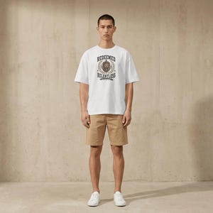 Puede incluir: Una camiseta blanca de manga corta con un gráfico central que muestra una cabeza de león, espigas de trigo y el texto "REDEEMED" arriba y "RELENTLESS" abajo, combinada con pantalones cortos tipo chino color canela y zapatillas blancas.