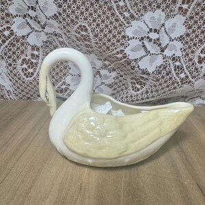 May include: A ceramic swan-shaped planter or decorative bowl featuring a glossy off-white finish with pale yellow accents on the wings and neck. The swan has a curved neck, a defined beak, and a hollow center designed for holding small items.