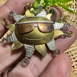 Puede incluir: Un broche con forma de sol hecho de metales mixtos, con una cara de latón con una sonrisa grabada y gafas de sol de metal color cobre. Los rayos del sol alternan entre plata texturizada y latón, acentuados con espirales de alambre y cuentas.