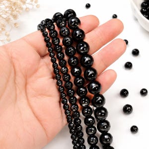 May include: Four strands of smooth, round, polished black beads arranged in increasing size order. The beads have a high-gloss finish that reflects light, creating bright white highlights on their surfaces. Several loose black beads are scattered nearby.