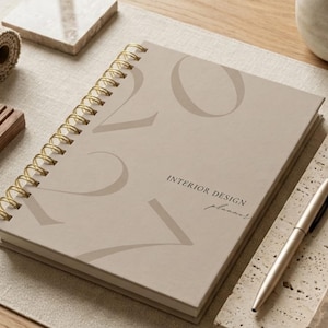 May include: A beige spiral-bound planner with gold-toned wire binding. The cover features the large, faint text 2024 and the printed words INTERIOR DESIGN planner in a dark font. A matching gold-toned ballpoint pen rests beside the planner.