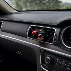 Peut inclure: Un désodorisant de voiture en forme de nez d'avion de chasse avec une hélice rouge, un corps noir et un motif de gueule de requin blanche aux dents acérées, fixé sur les lamelles horizontales d'une bouche d'aération de tableau de bord.