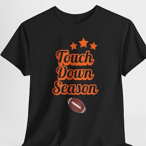Op de afbeelding: Een zwart t-shirt met korte mouwen voorzien van de oranje tekst Touch Down Season in een cursief lettertype, drie oranje sterren boven de tekst en een afbeelding van een bruine en witte football onder de tekst op de voorzijde.