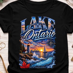 Può includere: Una t-shirt nera con una grafica del lago Ontario. Il design include un faro, una barca a vela, una rosa dei venti, una foglia d'acero rossa e la sagoma dello skyline sotto un cielo al tramonto. Il testo riporta Lake Ontario e Great Lakes.