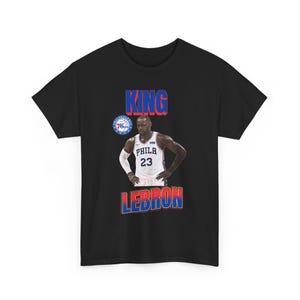 Può includere: Una t-shirt nera a maniche corte con la grafica di un giocatore di basket che indossa una maglia bianca dei Philadelphia 76ers con il numero 23. Il testo KING è stampato in blu sopra il giocatore e LEBRON in blu e rosso sotto.
