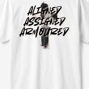 Pode incluir: Uma t-shirt branca de manga curta com um gráfico de cruz preta com pinceladas nas costas. Sobreposta à cruz está o texto preto ALIGNED ASSIGNED ARMOURED numa fonte a negrito de estilo manuscrito. O design está centrado na parte superior.