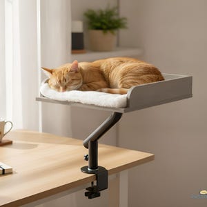 Può includere: Un gatto arancione che dorme su un cuscinetto bianco morbido sopra una piattaforma rettangolare grigia. La piattaforma è sostenuta da un braccio metallico nero regolabile con un morsetto fissato al bordo di una scrivania in legno. Il testo deskbeds è visibile.