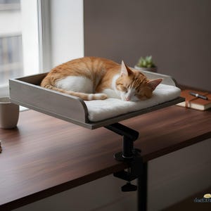 May include: A gray wooden cat bed platform with a white plush cushion, attached to a desk with a black metal C-clamp mount. An orange and white tabby cat is curled up sleeping on the soft white pad. The text deskbeds is in the corner.