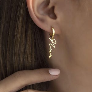 May include: A gold hoop earring featuring a dangling charm shaped like the word "Mum" in a cursive script font. The earring is made of polished gold metal and hangs from a small, circular hoop that secures through the earlobe.
