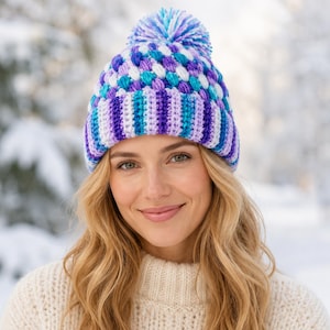 May include: A crochet beanie featuring a vibrant color palette of purple, blue, and white. The hat has a wide, vertically striped brim and a textured, bobble-stitch crown topped with a matching multi-colored yarn pom-pom for a winter look.