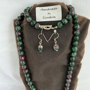 May include: A beaded necklace and matching drop earrings made of faceted green and magenta zoisite stones. The necklace features a silver-toned toggle clasp. A white rectangular tag with the black text "Handmade in Cumbria" is attached.