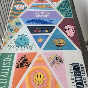 Op de afbeelding: Een rechthoekig tafeloppervlak beschilderd met een geometrisch patroon van driehoeken met diverse kleurrijke popcultuurillustraties, waaronder stripfiguren, smileys, bloemmotieven en de tekst Parental Advisory Explicit Content.
