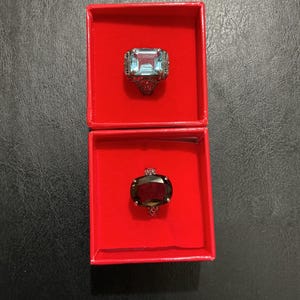 May include: Two rings displayed in separate red velvet-lined boxes. The top ring features a rectangular light blue gemstone set in a dark metal band with intricate filigree detailing. The bottom ring has a large oval dark red gemstone.