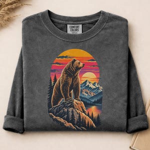 Puede incluir: Una camiseta de manga larga gris carbón con un estampado gráfico de un oso pardo de pie sobre un acantilado rocoso. El diseño incluye una puesta de sol sobre montañas, un lago y pinos. La etiqueta interior del cuello muestra el texto Comfort Colors.
