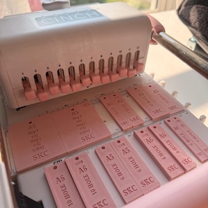 Op de afbeelding: Een lichtroze en witte Cinch-inbindmachine met een bovenste rij van twaalf verstelbare roze ponshendels en een basisplaat met gedrukte gidsen voor A5- en A6-papierformaten, inclusief labels voor het aantal gaten en SKC-merktekens.