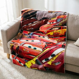 May include: A soft plush throw blanket featuring a repeating graphic print of the red race car character Lightning McQueen from the movie Cars. The design displays multiple angles of the car with the number 95 in yellow and orange accents.