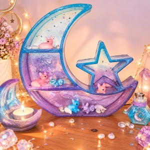 May include: A crescent moon and star-shaped display shelf featuring a gradient of blue, purple, and pink resin with embedded glitter. The shelf contains small animal figurines, including foxes and rabbits, in various pastel colours and finishes.