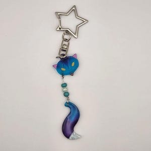 Peut inclure: Un porte-clés composé d'un fermoir en forme d'étoile argentée attaché à une breloque tête de renard bleue et violette, suivie de deux perles turquoise et une perle blanche, se terminant par une queue de renard courbée dégradée bleu et violet.