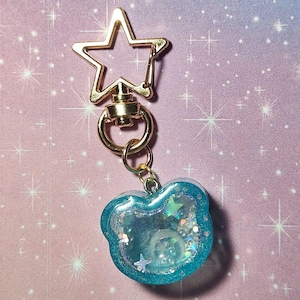 May include: A gold-toned star-shaped keychain clasp attached to a circular ring and a blue bear-shaped shaker charm. The charm features a translucent blue resin border filled with liquid, small white beads, and iridescent star-shaped glitter.