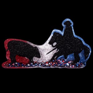May include: A decorative resin ornament featuring a black silhouette of a person on horseback and a bull. The design incorporates red, white, and blue glitter accents, with a clear border outlining the shapes against a solid black background.